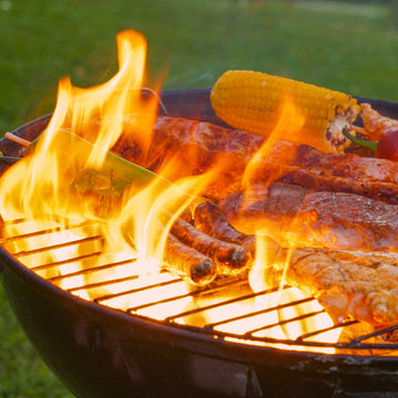 High flames flare up through the metal grates of an outdoor charcoal grill loaded with sausages, meat cuts, and a cob of corn.