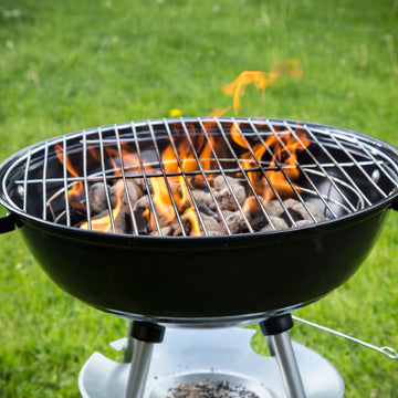 Black round kettle charcoal grill with open flames rising through the metal cooking grate over burning charcoal briquettes outdoors on a green lawn.