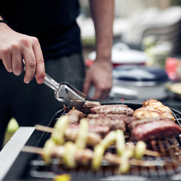 Close-up of a person using metal tongs to flip sausages, meats, and vegetable skewers on a portable electric outdoor grill.
